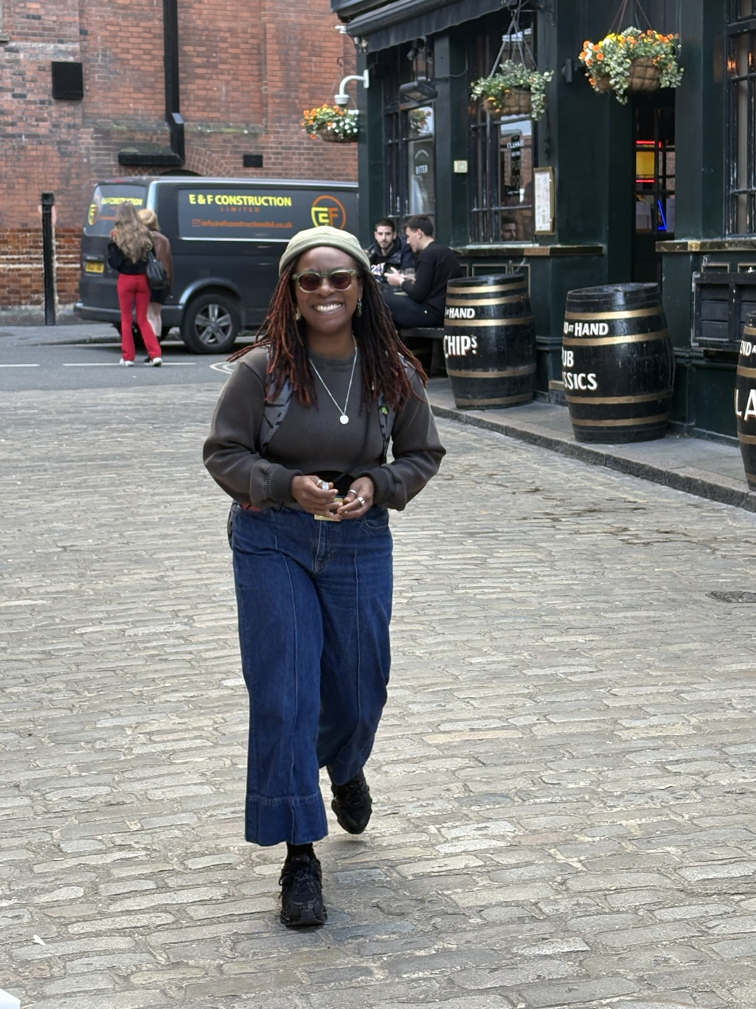 A woman with long, curly hair and sunglasses walking towards the camera on a cobblestone street, smiling. She is wearing a grey sweater and wide-legged blue jeans. In the background, there is a pub with hanging flower baskets and a construction vehicle.